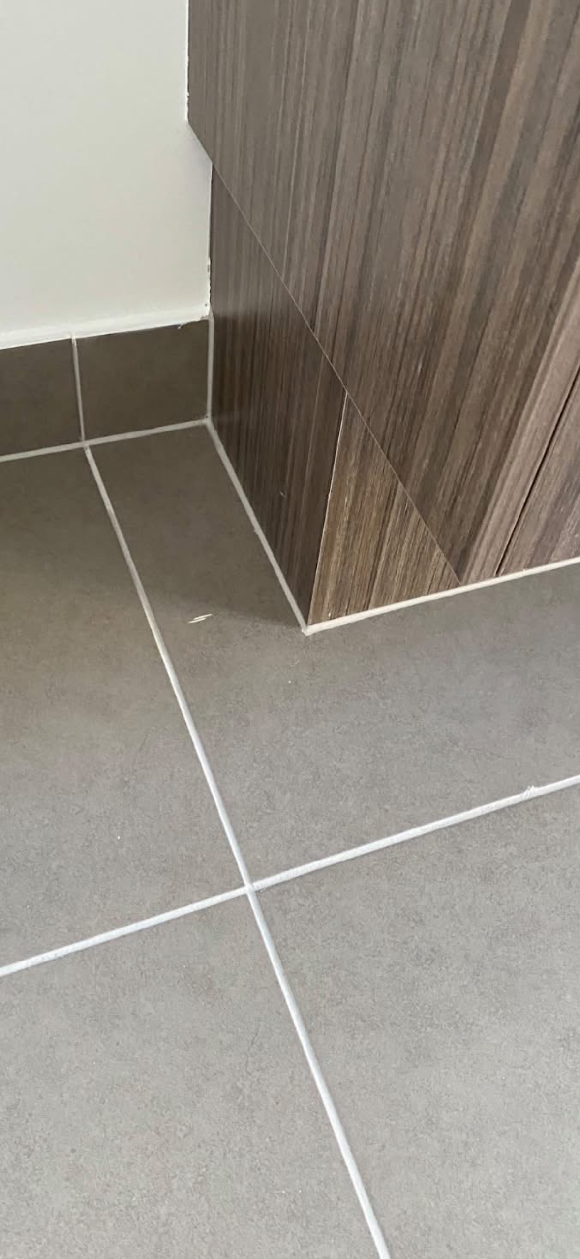 A grey tiled floor meeting a timber look kitchen kickboard, with a chip visible in one floor tile near the corner of the cabinetry.