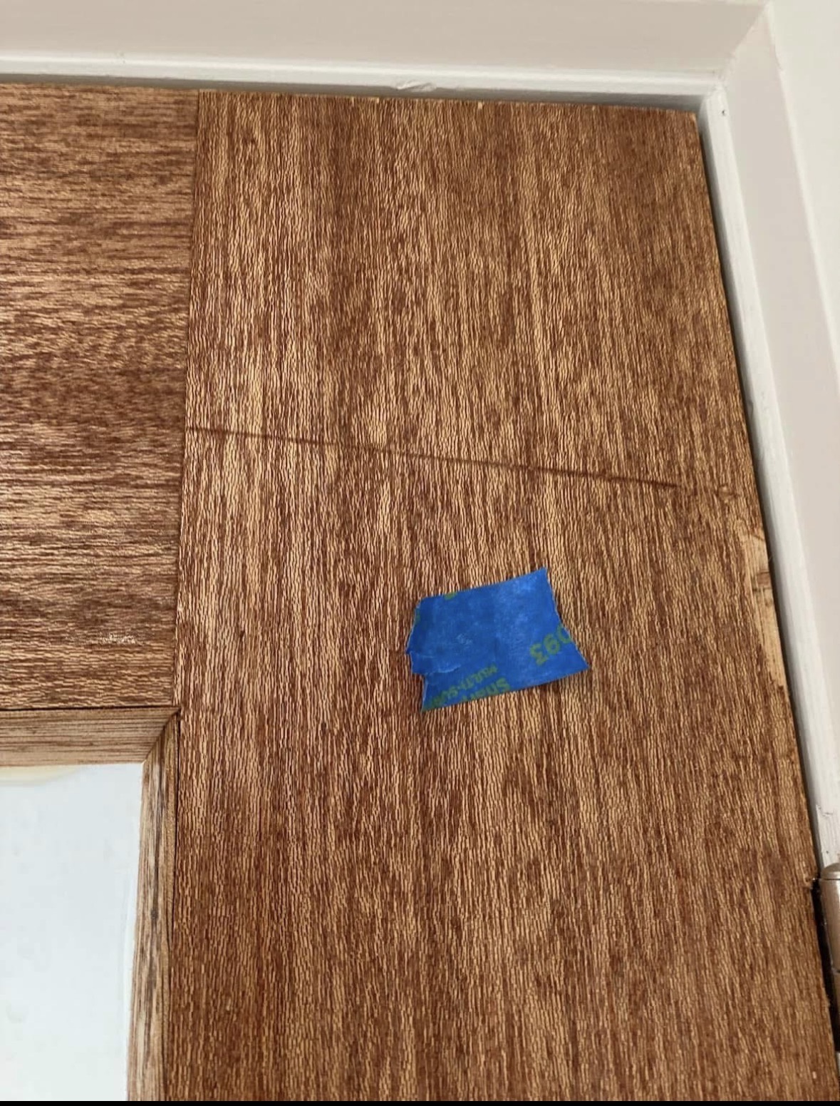 A stained timber front door photographed up close, with a strip of blue tape marking damage to the timber surface beside the door frame.