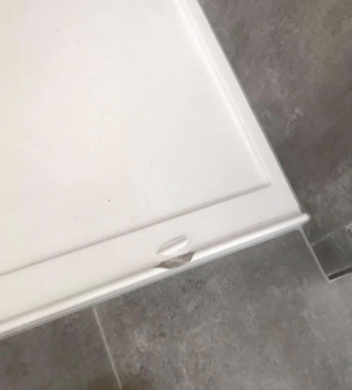 The corner of a white shower base with a chip broken out of its moulded edge, showing grey material under the white surface, against grey floor tiles.