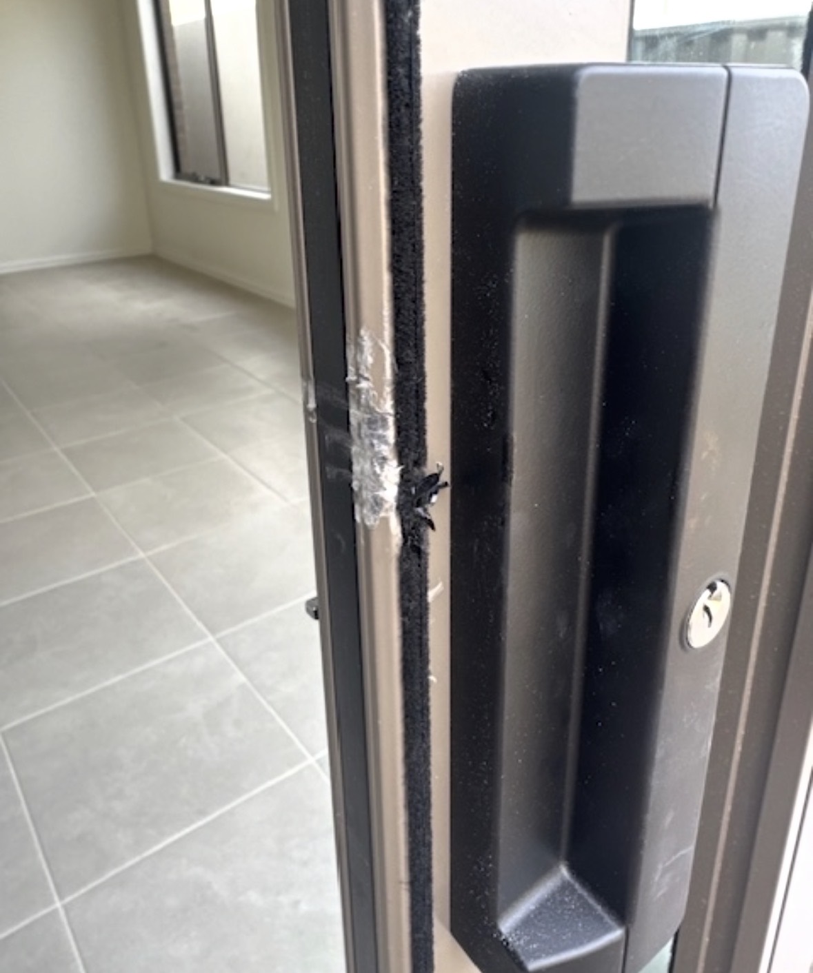 A black aluminium sliding door frame beside the handle, with metal damaged in an attempted break in and clear tape over the damage near the lock keeper.