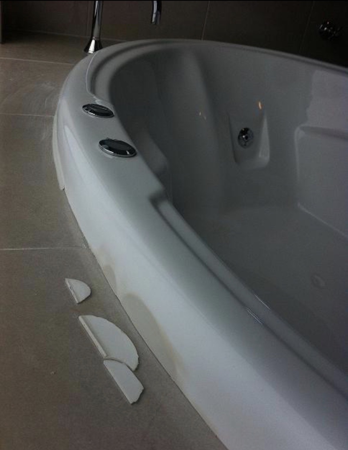 A white corner spa bath with a large piece broken out of its rim, and three broken shards of white acrylic lying on the tiled floor beside it.