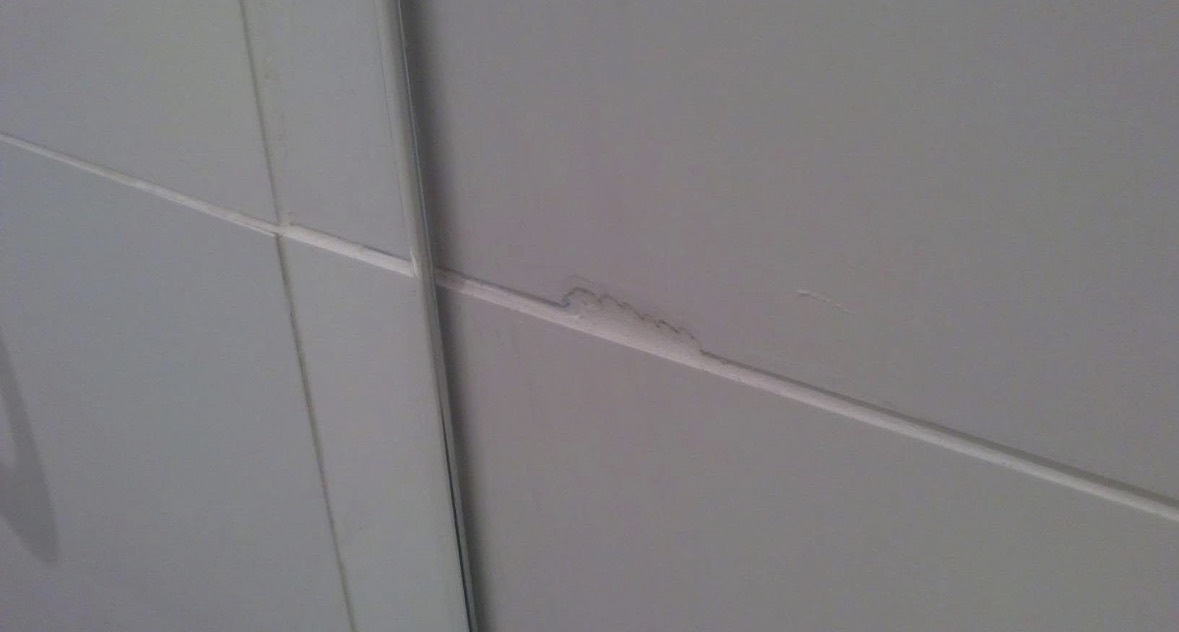 A pale grey ceramic wall tile with a chip broken out of its lower edge where it meets the grout line, exposing the rough body of the tile underneath the glaze.