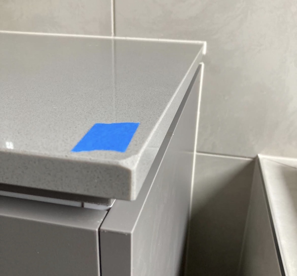 A grey engineered stone vanity top photographed at the corner, with a square of blue masking tape on the surface marking a chip broken out of the front edge below it.